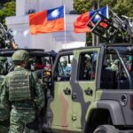 Soldiers in camouflage uniforms standing near military vehicles with flags in the background