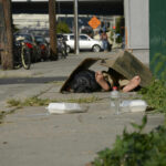 A person lying on the sidewalk under a cardboard box in an urban setting