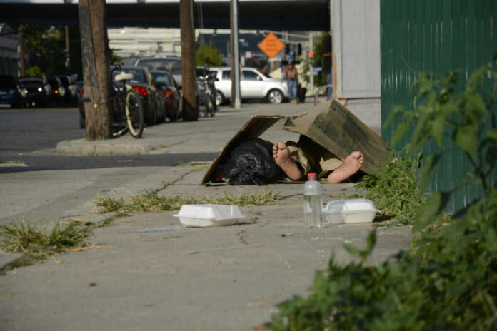 shutterstock_716983525.jpg A person lying on the sidewalk under a cardboard box in an urban setting