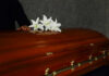A wooden casket adorned with white lilies, with a hand resting on it