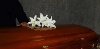 A wooden casket adorned with white lilies, with a hand resting on it