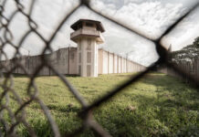 View of a prison tower surrounded by a security fence and grassy area under a cloudy sky