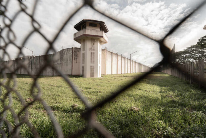 shutterstock_1587239341.jpg View of a prison tower surrounded by a security fence and grassy area under a cloudy sky