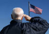 Elderly man saluting in front of an American flag against a blue sky