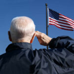 Elderly man saluting in front of an American flag against a blue sky