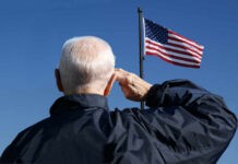 Elderly man saluting in front of an American flag against a blue sky