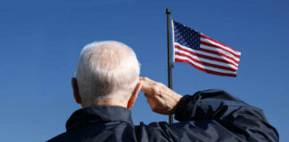 Elderly man saluting in front of an American flag against a blue sky