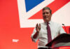 A man in a white shirt and tie gestures while speaking at a podium with a British flag backdrop