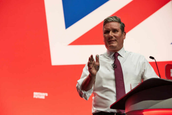 A man in a white shirt and tie gestures while speaking at a podium with a British flag backdrop