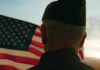 A veteran in uniform standing in front of an American flag during sunset