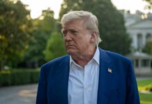 A political figure with a serious expression standing outdoors near the White House