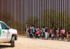 Group of families and children walking near a border wall with a customs vehicle nearby