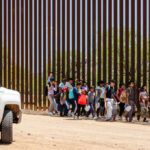 Group of families and children walking near a border wall with a customs vehicle nearby