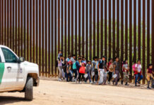 Group of families and children walking near a border wall with a customs vehicle nearby