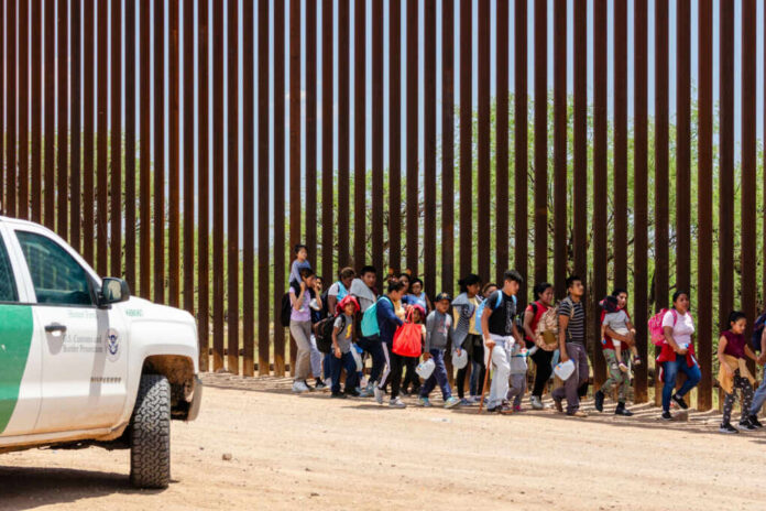 shutterstock2333258555jpg Group of families and children walking near a border wall with a customs vehicle nearby