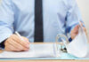 A person in a blue shirt writing on a document while holding a binder