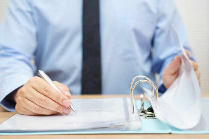 A person in a blue shirt writing on a document while holding a binder