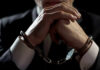 A man in a suit with hands clasped, wearing handcuffs against a dark background