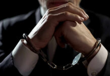 A man in a suit with hands clasped, wearing handcuffs against a dark background