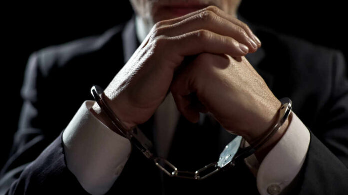 A man in a suit with hands clasped, wearing handcuffs against a dark background