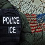 Death Threats Target Agents’ Families A police officer in a tactical vest stands near a barbed wire fence with an American flag in the background