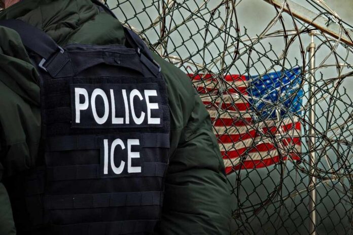 An,Immigration,And,Customs,Enforcement,(ice),Officer,Stands,In,Front A police officer in a tactical vest stands near a barbed wire fence with an American flag in the background
