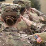 Female soldier saluting during a military ceremony with fellow soldiers in camouflage uniforms