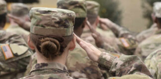 Female soldier saluting during a military ceremony with fellow soldiers in camouflage uniforms