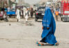 A woman in a blue burqa walking on a dusty street with pedestrians and vehicles in the background