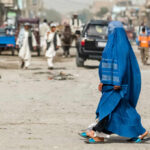 A woman in a blue burqa walking on a dusty street with pedestrians and vehicles in the background