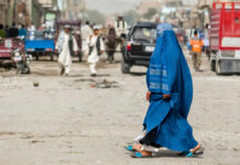 A woman in a blue burqa walking on a dusty street with pedestrians and vehicles in the background