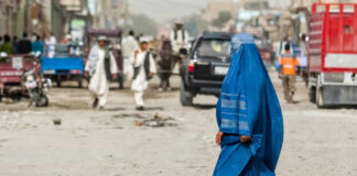 A woman in a blue burqa walking on a dusty street with pedestrians and vehicles in the background