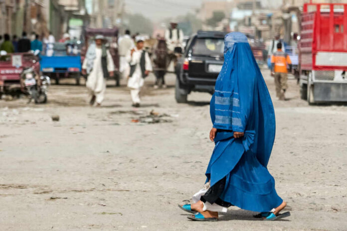 shutterstock2196739241jpg A woman in a blue burqa walking on a dusty street with pedestrians and vehicles in the background