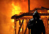 A hooded figure holding a lighter in front of a burning structure