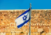 Ceasefire Shakes After Generals Killed Israeli flag waving in front of a stone wall under a clear blue sky