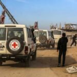 Red Cross vehicles drive through a dusty street with damaged buildings