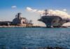 Firestorms Cripple $13B Supercarriers Aircraft carrier and support ships docked at a naval harbor