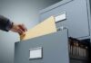 Hand filing a manila folder into an open metal filing cabinet drawer