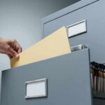 Hand filing a manila folder into an open metal filing cabinet drawer