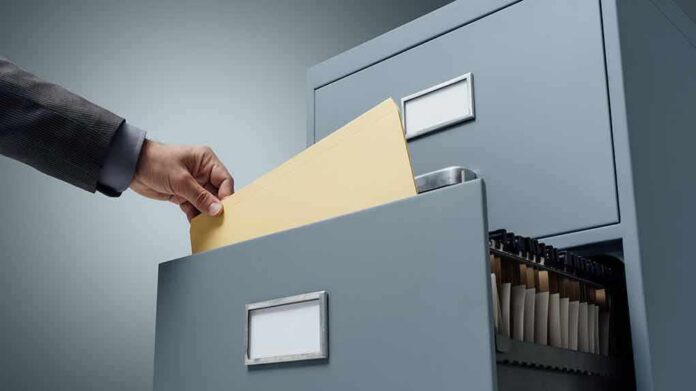 Hand filing a manila folder into an open metal filing cabinet drawer
