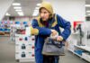 Person in hooded jacket placing item into a gray bag in a store aisle