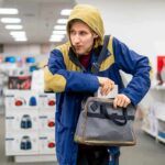 Person in hooded jacket placing item into a gray bag in a store aisle