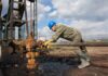 Worker in blue hard hat operating rusty oil pump equipment outdoors