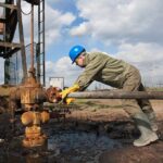 Worker in blue hard hat operating rusty oil pump equipment outdoors