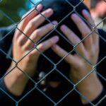 Person behind chain-link fence holding head in hands