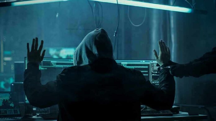 Hooded person at computer raises hands as another clamps handcuffs
