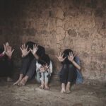 group of children sitting against a cracked wall with hands raised to shield faces