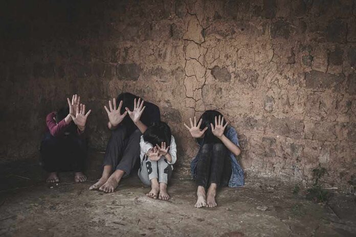 group of children sitting against a cracked wall with hands raised to shield faces